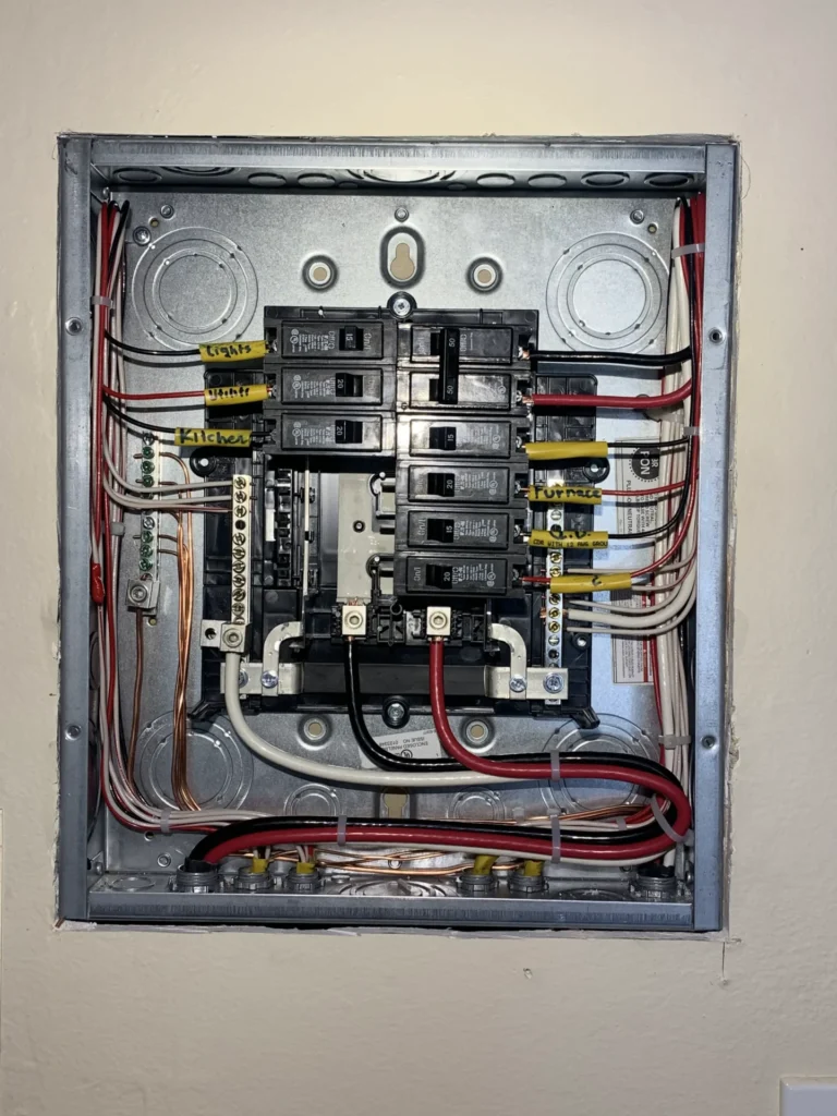 An open electrical panel with multiple circuit breakers, wiring, and labels indicating different circuits for various parts of a building. The panel is mounted on a beige wall.