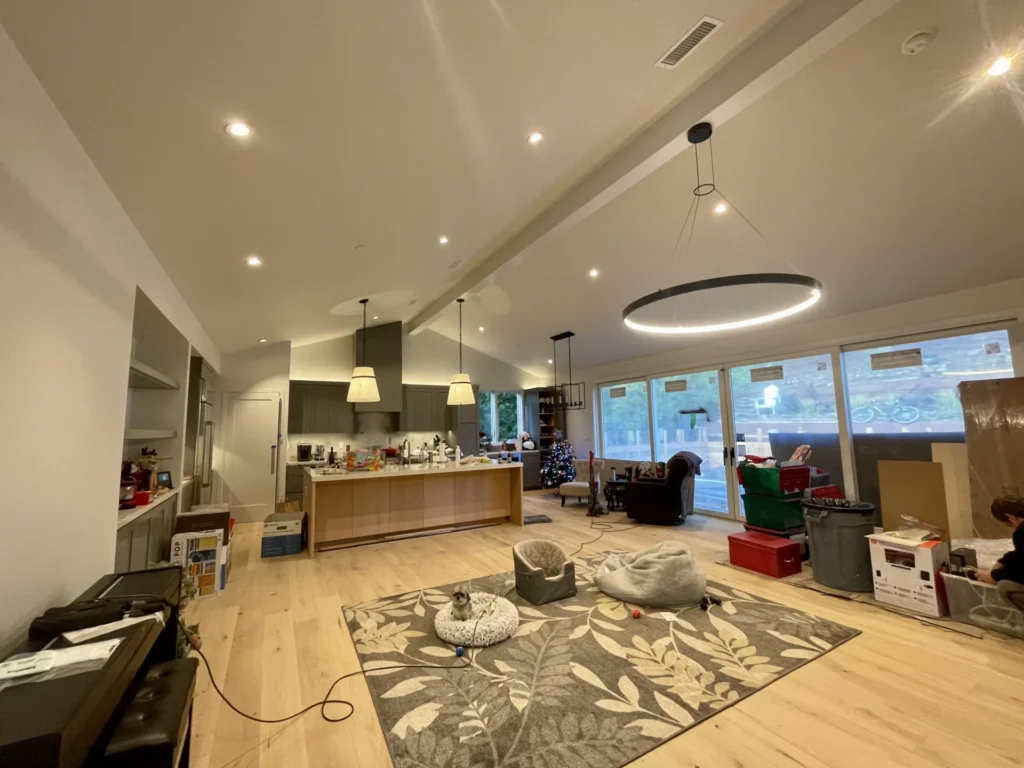 A spacious living area with a high ceiling, modern lighting fixtures, a kitchen island, and large windows. There's a cluttered counter, scattered boxes, and a decorated Christmas tree in the corner. Sf Electrician