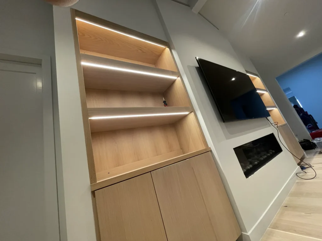 A modern living room with built-in wooden shelves with minimalist LED lighting, a flat-screen TV mounted on the wall, and a sleek fireplace below the TV.