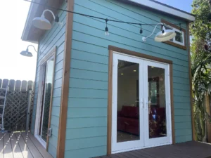 A small, teal, wooden house with double glass doors and string lights hanging above. The interior is partially visible, showing a red couch.