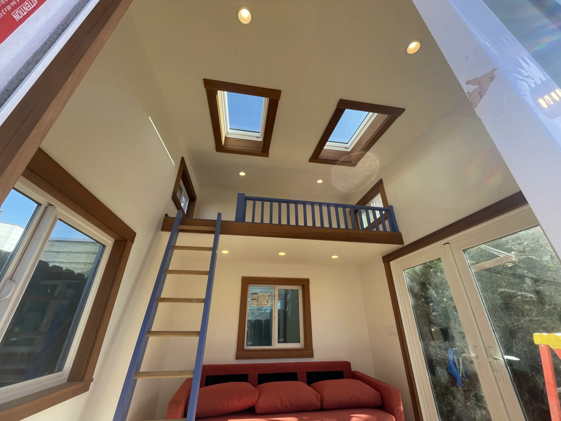 Interior of a tiny house featuring a loft with skylights, accessible by a ladder. Below, a red couch sits under the loft railing, adjacent to glass doors.