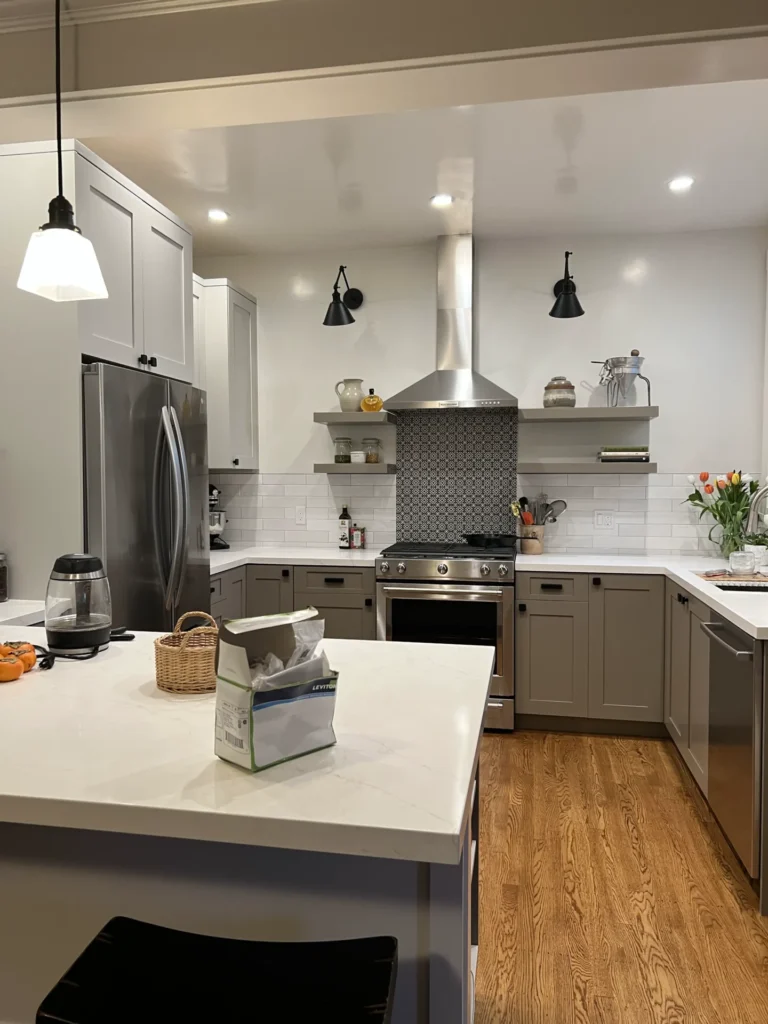 A modern kitchen with grey cabinets, stainless steel appliances, a central island, and a wooden floor. The countertop has various kitchen items and a bag of flour. Ceiling lights and under-cabinet lights are on. remodel electrical