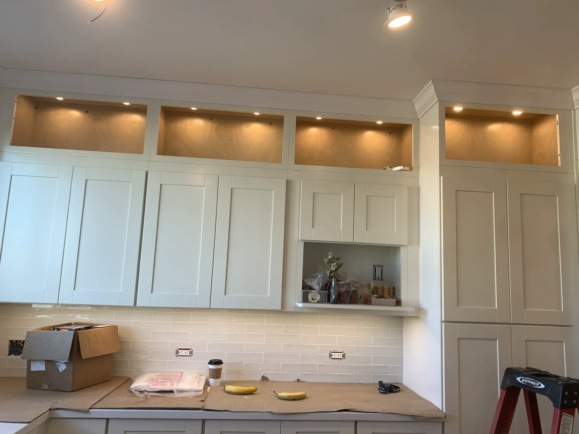 Kitchen under renovation with white cabinets and newly installed recessed lights. Unfinished cupboards, boxes, and tools are scattered on the countertop and floor.