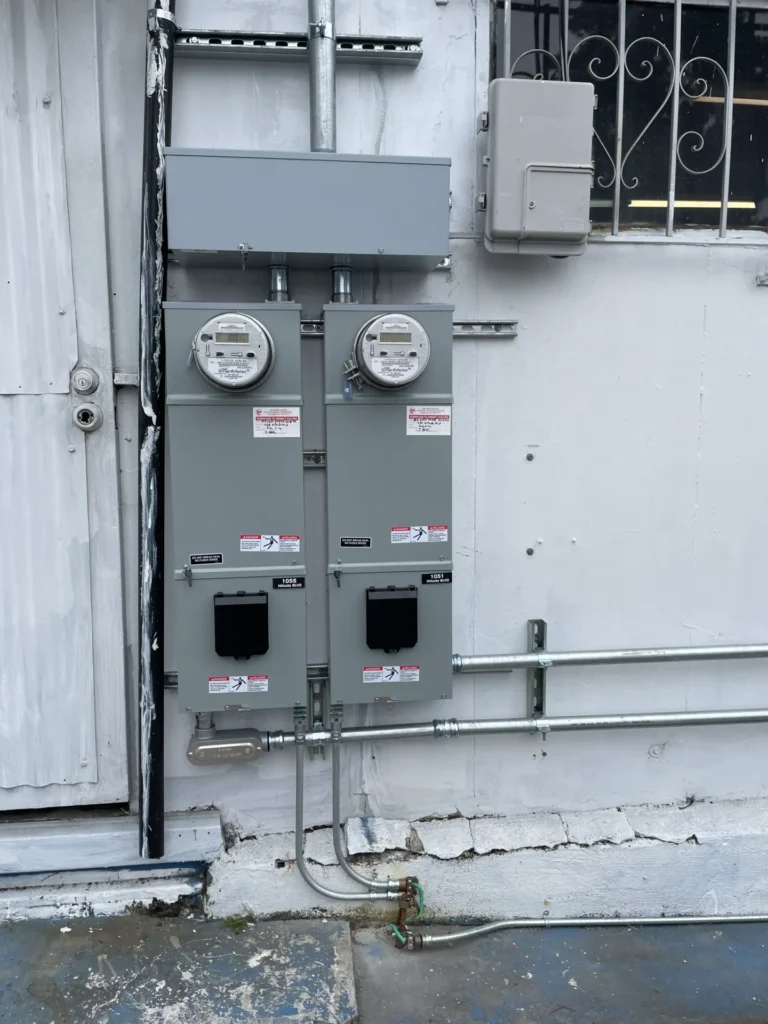 Two electrical meter boxes mounted on an exterior wall, with conduit pipes running below and to the side. Nearby is a door with peeling paint and a barred window.