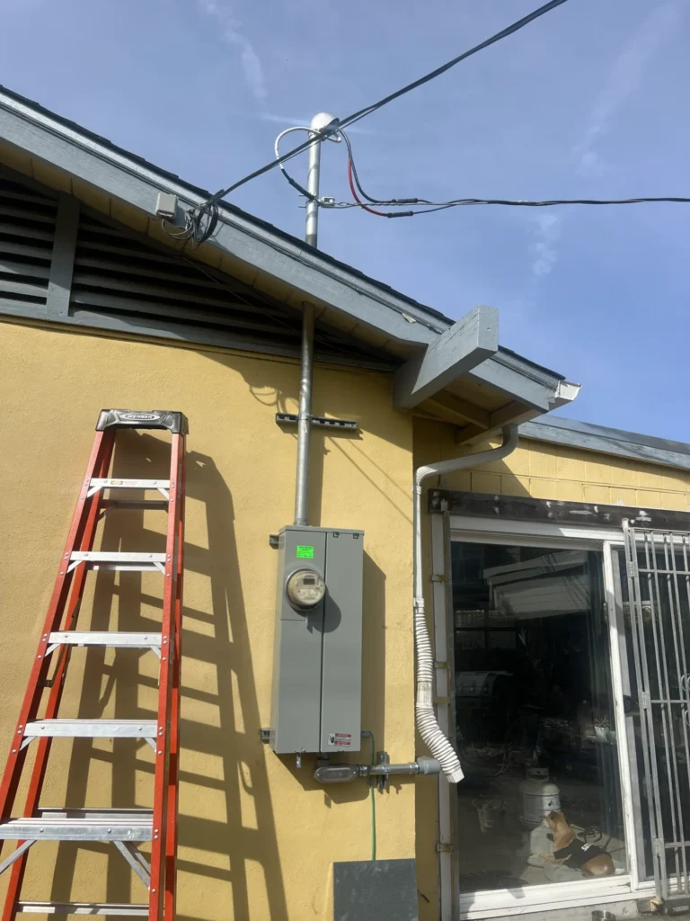 Red ladder leaning against a yellow building beside an electrical panel, with wiring extending to the roof and a sliding glass door nearby.